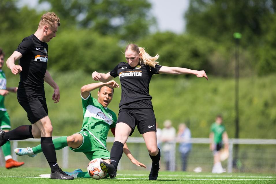 Drei Fußballspieler in Aktion auf einem Rasenplatz, eine Spielerin in Schwarz springt über einen am Boden liegenden Spieler.