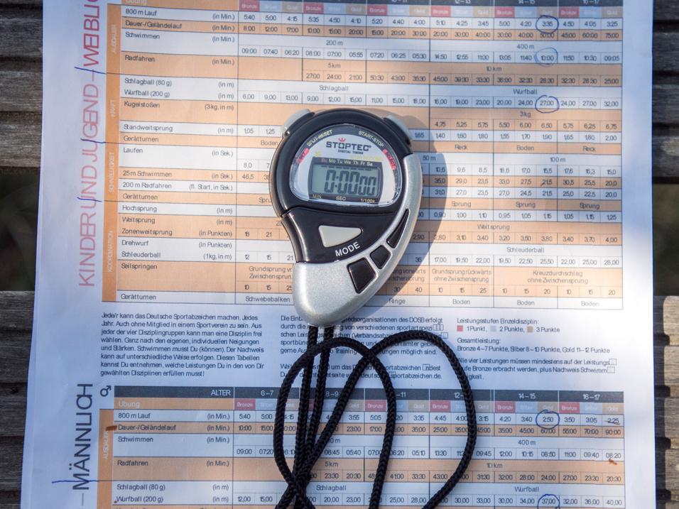 Stoppuhr und Infoblatt des Deutschen Sportabzeichens auf einer Holzbank im Freien.